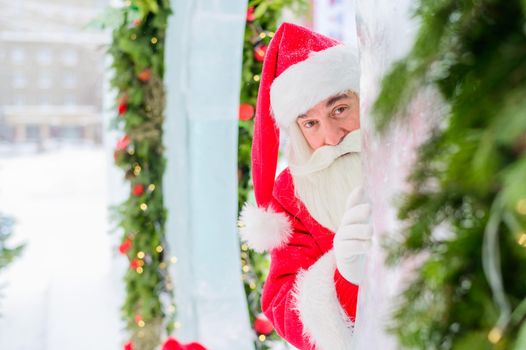 Santa claus peeks out of Christmas decorations outdoors.