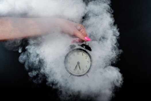 A woman holds an alarm clock in a studio full of smoke. White fog enveloped a round retro mechanical watch