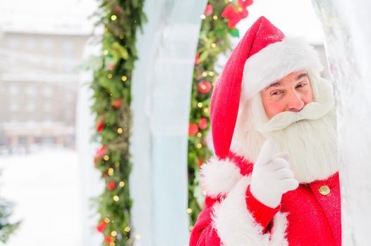 Santa claus peeks out of Christmas decorations outdoors.