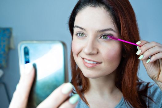 Beautiful redhead woman paints eyelashes and takes a selfie. The girl puts mascara and looks at the phone instead of a mirror