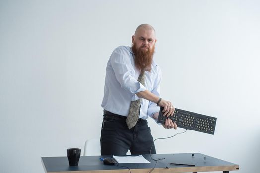 Angry bald man with a red beard in the office in a business suit crashes a computer. Manager with a nervous breakdown breaks the keyboard on the monitor