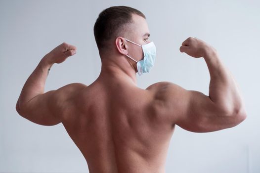 Male bodybuilder in a medical mask stands with his back. The guy goes in for sports in quarantine. Respiratory protection. Shows biceps