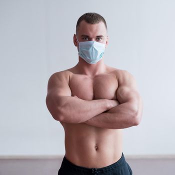 A male bodybuilder in a medical mask crossed his arms over his muscular bare chest. The guy goes in for sports in quarantine. Respiratory protection