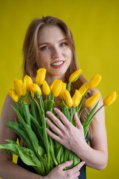 Caucasian woman with an armful of yellow tulips on a yellow background. International Women's Day. Bouquet of spring flowers.