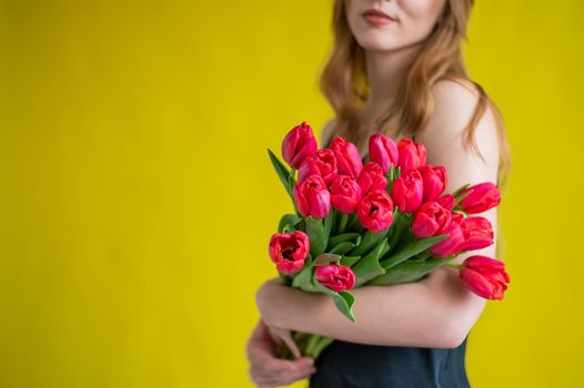 Unrecognizable woman with a bouquet of red tulips on a yellow background. A girl in a black dress holds an armful of flowers. A gift for International Women's Day. Spring holiday