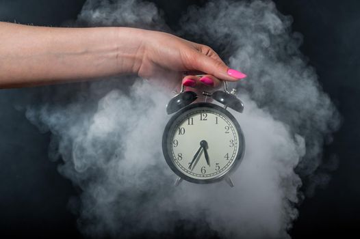 A woman holds an alarm clock in a studio full of smoke. White fog enveloped a round retro mechanical watch