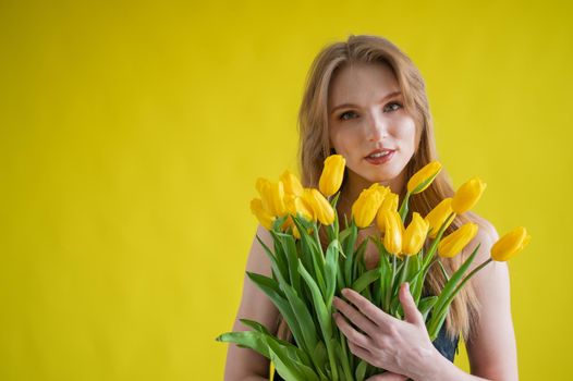 Caucasian woman with an armful of yellow tulips on a yellow background. International Women's Day. Bouquet of spring flowers.