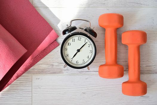 Top view on orange dumbbell alarm clock and raspberry yoga mat. The concept of home sports during quarantine. Fitness time. Early rise for classes