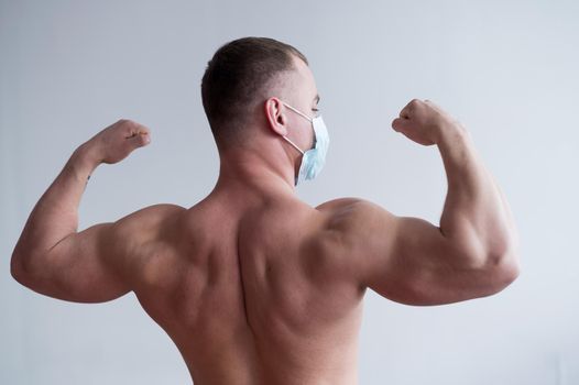 Male bodybuilder in a medical mask stands with his back. The guy goes in for sports in quarantine. Respiratory protection. Shows biceps