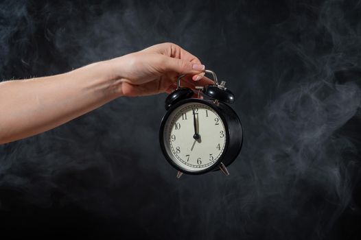 Close-up of a female hand holding a clock on a black background in smoke. Alarm clock at midnight in a mystical fog