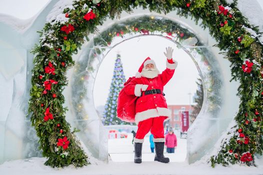 Santa Claus posing with a bag of gifts on the background of Christmas decorations outdoors.