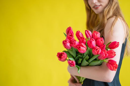Unrecognizable woman with a bouquet of red tulips on a yellow background. A girl in a black dress holds an armful of flowers. A gift for International Women's Day. Spring holiday