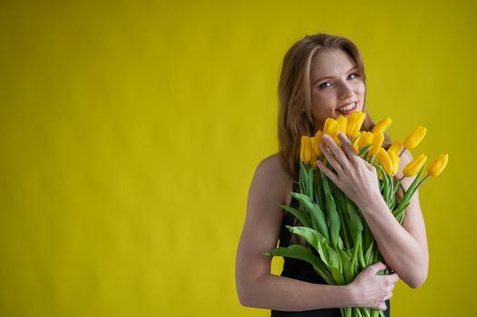 Caucasian woman with an armful of yellow tulips on a yellow background. International Women's Day. Bouquet of spring flowers.