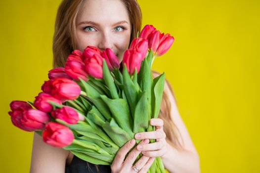 Woman with a bouquet of red tulips on a yellow background. Happy girl in a black dress holds an armful of flowers. Gift for Valentine's Day. The most romantic day of the year