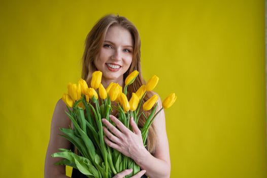 Caucasian woman with an armful of yellow tulips on a yellow background. International Women's Day. Bouquet of spring flowers.