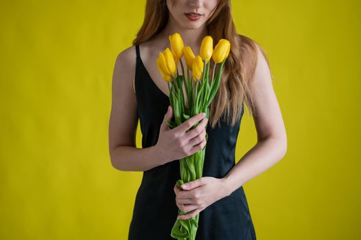 Unrecognizable woman with a bouquet of tulips on a yellow background. A girl in a black dress holds an armful of flowers. A gift for International Women's Day. Spring holiday