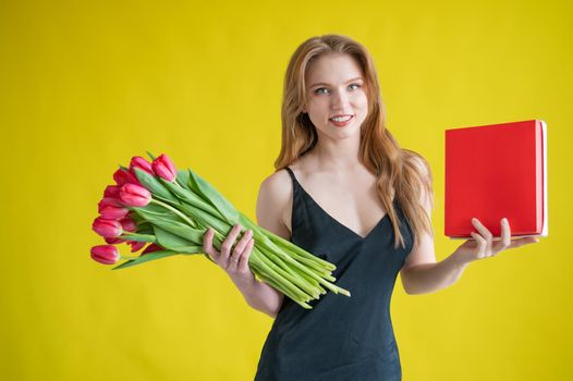 Woman with a bouquet of red tulips on a yellow background. Happy girl in a black dress holds an armful of flowers and a box of chocolates. Gift for Valentine's Day. The most romantic day of the year