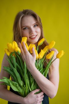 Caucasian woman with an armful of yellow tulips on a yellow background. International Women's Day. Bouquet of spring flowers.