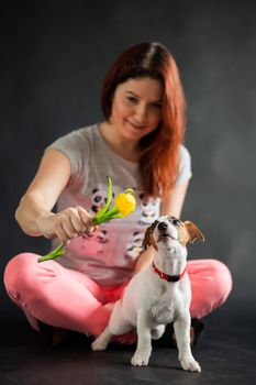 Red-haired woman holding a yellow tulip and playing with her puppy on a black background.