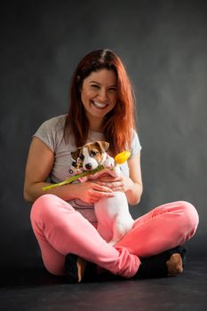 Happy smiling red-haired woman holding a small funny puppy with a yellow tulip in it mouth. The joyful owner plays with the dog Jack Russell Terrier on a black background.
