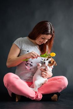 Red-haired woman holding a yellow tulip and playing with her puppy on a black background.