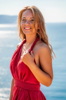 Smiling young woman in a red dress looks at the camera. A beautiful tanned girl enjoys her summer holidays at the sea. Portrait of a stylish carefree woman laughing at the ocean