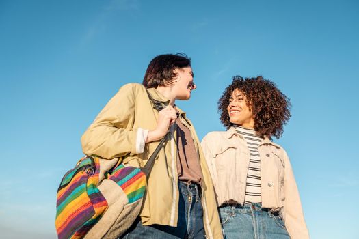 two young women smiling and looking at each other against a blue sky background, concept of female friendship and racial diversity