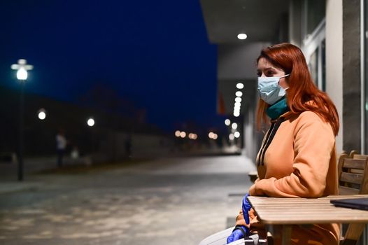 A woman in a medical mask and gloves walks late at night alone. The girl is sitting at a wooden table with a laptop in quarantine. Coronavirus