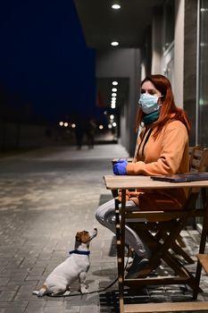 A woman in a medical mask and gloves walks late at night with her little dog. A girl sits on a chair in the street with a Jack Russell Terrier in quarantine. Coronavirus