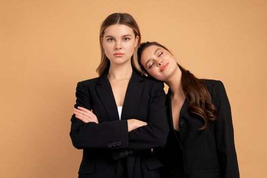 Two young business woman dressed black suit standing studio orange color background. One woman looking looking at camera arms crossed on chest other woman put her head on her shoulder. Friends support