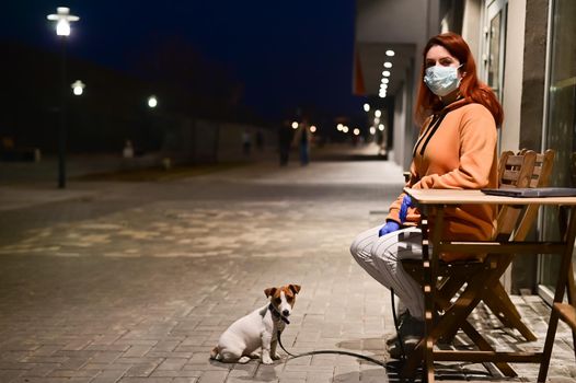 A woman in a medical mask and gloves walks late at night with her little dog. A girl sits on a chair in the street with a Jack Russell Terrier in quarantine. Coronavirus