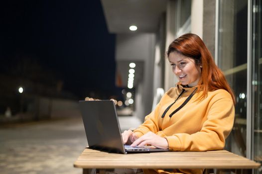 A woman works remotely at a laptop in a summer cafe late in the evening. A smiling girl is studying while sitting down in a wasted street at a wooden table