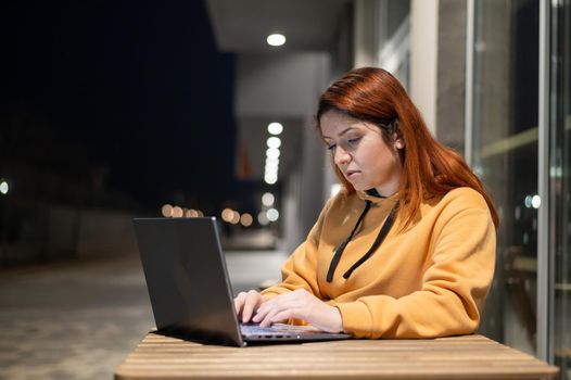 A woman works remotely at a laptop in a summer cafe late in the evening. Serious Girl studying while sitting on an empty street at a wooden table. Female freelancer in a sweatshirt