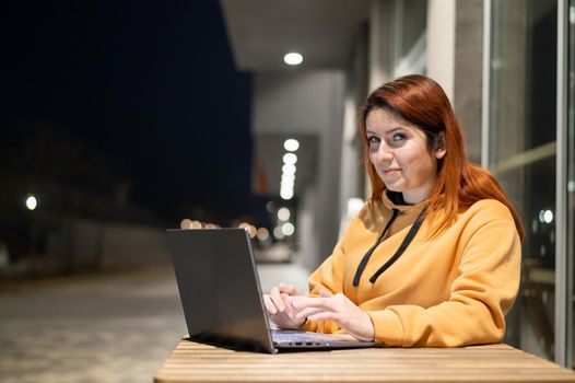 A woman works remotely at a laptop in a summer cafe late in the evening. A smiling girl is studying while sitting down in a wasted street at a wooden table