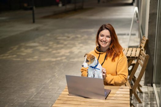 Smiling woman working on laptop at a wooden table in the street. The girl looks at the monitor with her pet jack russell terrier. Freelancer walks the dog in the evening