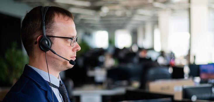 Portrait of a male call center operator in headset at workplace. A man works in an open space office answering customer calls. Support service or hotline. Agent of a telemarketing company