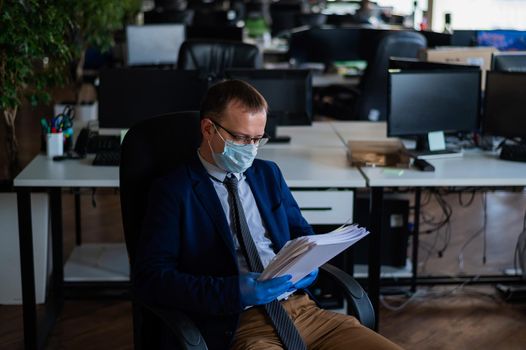 A man in a business suit and medical mask reads a paper report in an empty open space office. Social distance and isolation of employees. Urgent work during quarantine
