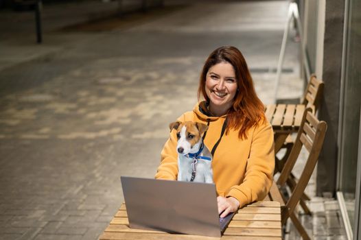 Smiling woman working on laptop at a wooden table in the street. The girl looks at the monitor with her pet jack russell terrier. Freelancer walks the dog in the evening