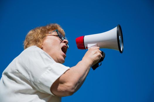 Emotional senior woman makes a speech in a megaphone on the outside. A pensioner yells into a sound amplifier on a blue background. female leader of the rally voices the conditions in the loudspeaker