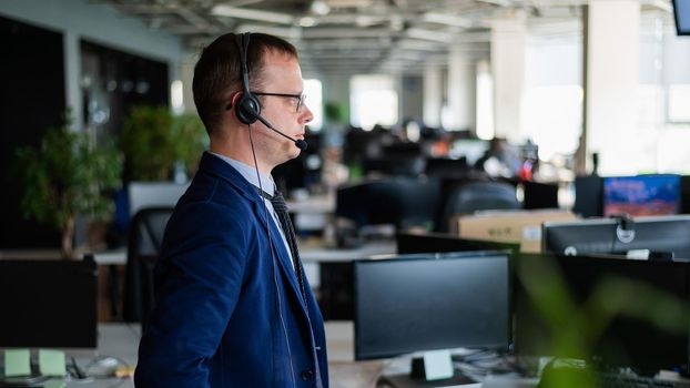 Portrait of a male call center operator in headset at workplace. A man works in an open space office answering customer calls. Support service or hotline. Agent of a telemarketing company