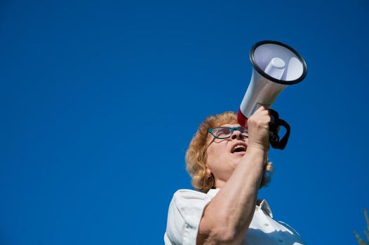 Emotional senior woman makes a speech in a megaphone on the outside. A pensioner yells into a sound amplifier on a blue background. female leader of the rally voices the conditions in the loudspeaker