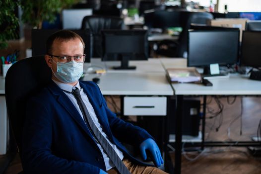 Serious man in a business suit and glasses in an empty office during quarantine. Male manager in a medical mask at the workplace. Social distance and workspace disinfection