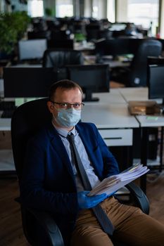 A man in a business suit and medical mask reads a paper report in an empty open space office. Social distance and isolation of employees. Urgent work during quarantine