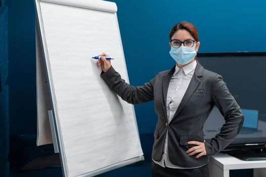 A woman in a medical mask leads a seminar in a conference hall. Business coach writes on a paper white board in the office. Female employee demonstrates presentation while maintaining social distance