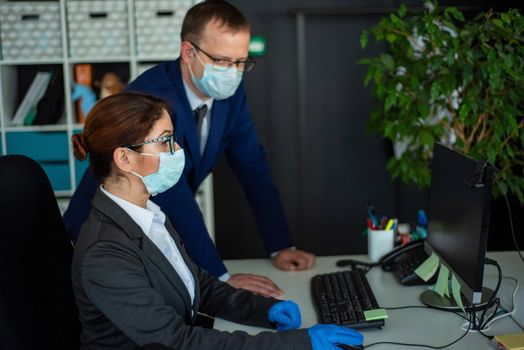 Colleagues in surgical masks in an open office space communicate at the work desk. A male top manager teaches a female intern how to work at a computer. The boss directs the subordinate