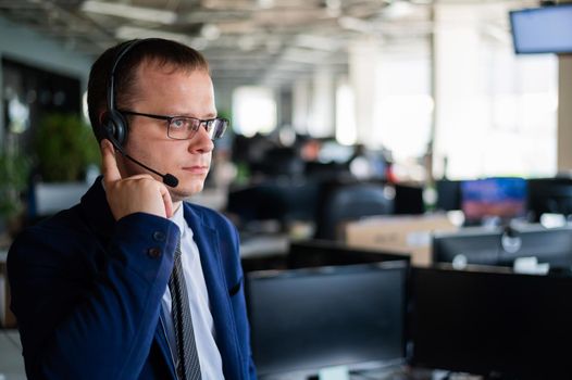 Portrait of a male call center operator in headset at workplace. A man works in an open space office answering customer calls. Support service or hotline. Agent of a telemarketing company