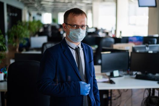 Serious man in a business suit and glasses in an empty office during quarantine. Male manager in a medical mask at the workplace. Social distance and workspace disinfection