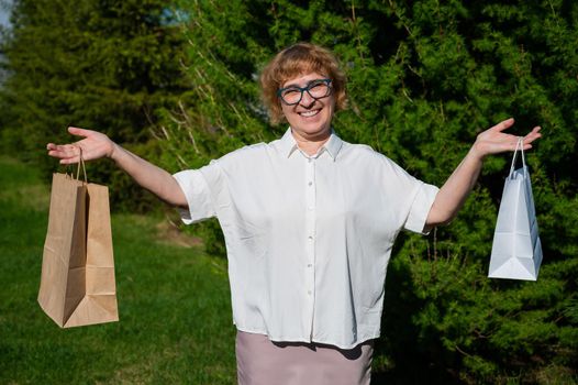 A happy elderly woman with glasses stands in a park and holds paper bags. Female retired shopaholic boasts of shopping. The concept of using eco friendly products to protect the environment