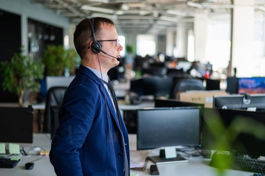 Portrait of a male call center operator in headset at workplace. A man works in an open space office answering customer calls. Support service or hotline. Agent of a telemarketing company