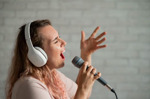 Close-up portrait of a caucasian woman with curly hair singing into a microphone. Beautiful emotional girl in white headphones sings a song in home karaoke and actively gestures against a brick wall
